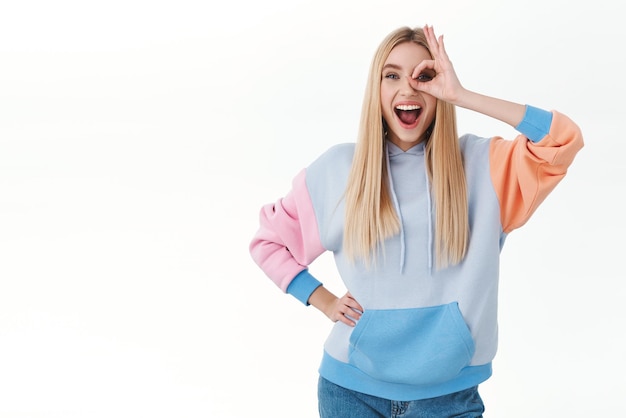 Menina loira sorridente, divertida e animada com cabelo comprido, mostrando sinal de bom sobre o olho e olhar otimista