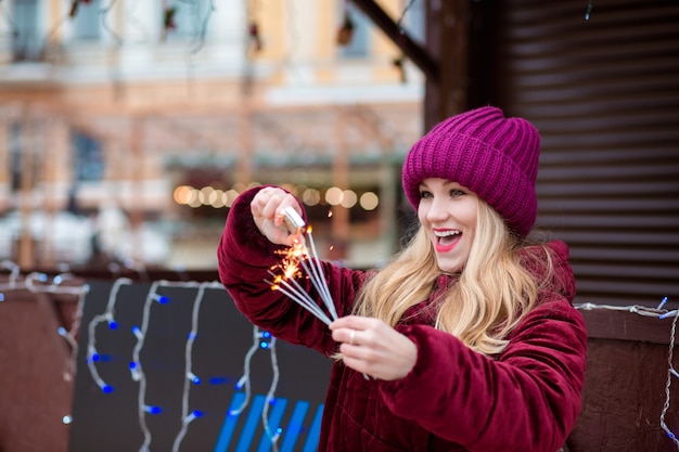 Menina loira surpresa vestida com roupas elegantes, ateia fogo a fogos de artifício brilhantes na feira de Natal em Kiev