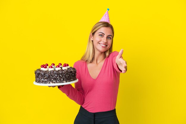 Menina loira uruguaia segurando bolo de aniversário isolado em fundo amarelo apertando as mãos para fechar um bom negócio