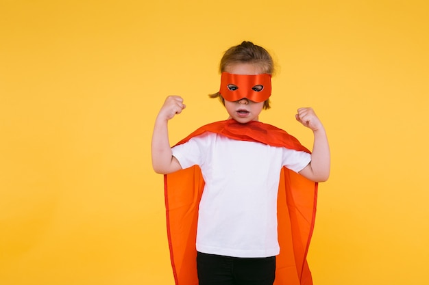 Menina loira vestida de super-heroína super-heroína com capa e máscara vermelhas, apertando os braços em sinal de força, sobre fundo amarelo