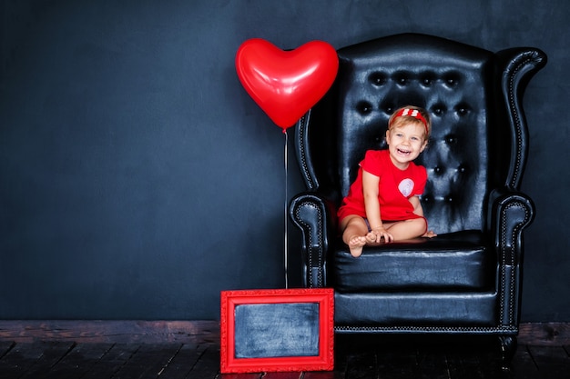 Menina loura pequena no vestido vermelho com a grinalda vermelha com os corações que sentam-se na poltrona com o balão vermelho do coração no dia do valentim do st.