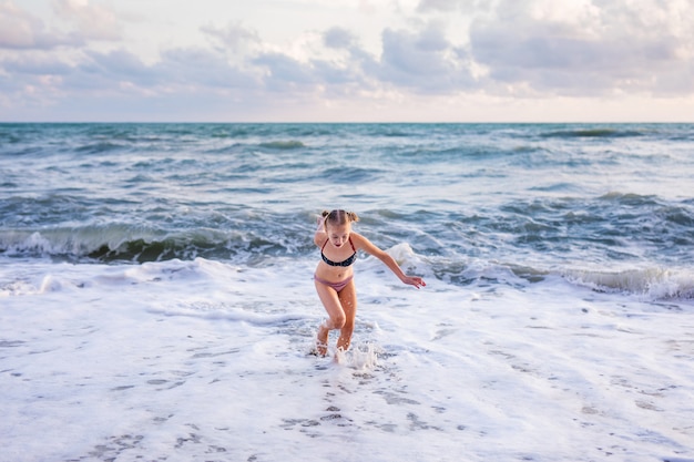 Menina loura que corre e que salta na praia na costa de mar azul em férias de verão no tempo do dia.