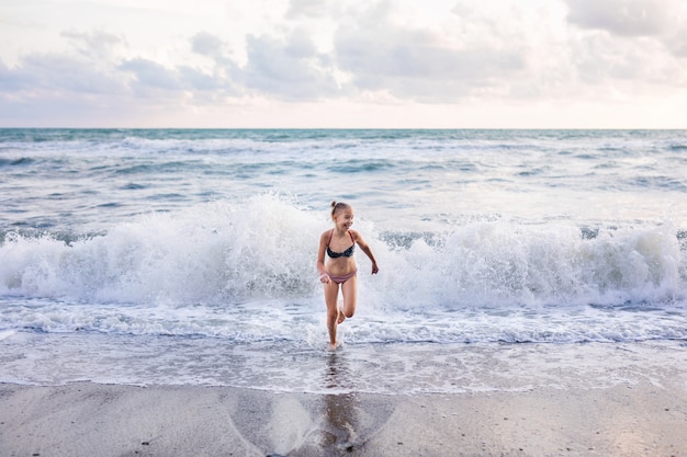 Menina loura que corre e que salta na praia na costa de mar azul em férias de verão no tempo do dia.