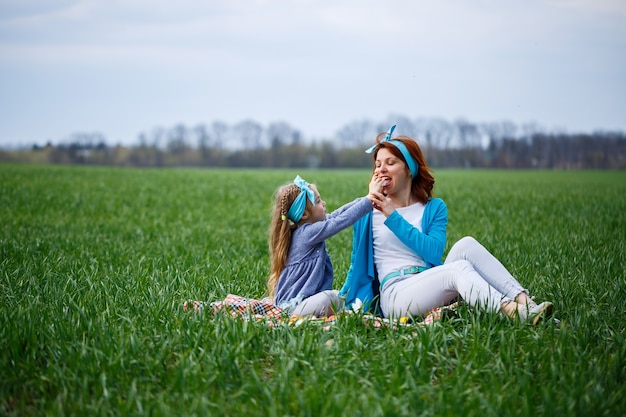 Menina menina e mãe mulher sentada na colcha e comendo biscoitos e geleia, grama verde no campo, clima ensolarado de primavera, sorriso e alegria da criança, céu azul com nuvens