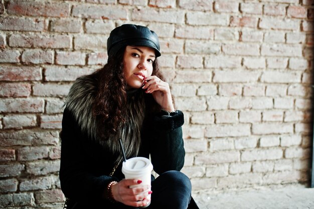 Menina mexicana encaracolada na tampa de couro e xícara de café plástica à mão andando nas ruas da cidade.
