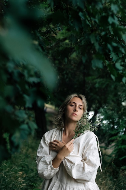 Menina misteriosa melancólica em um vestido branco fica com um buquê de flores brancas na floresta.