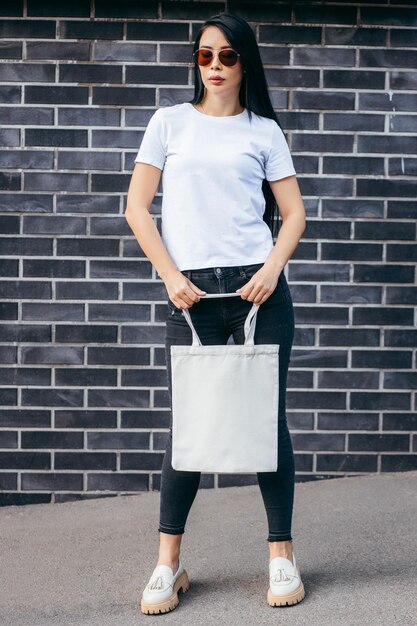 Menina modelo asiática jovem vestida com uma camiseta na rua segurando uma bolsa ecológica branca simulada