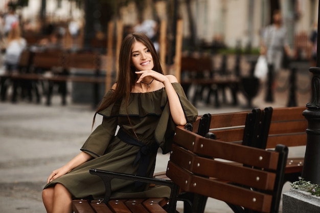 Menina modelo de vestido está sentado no banco marrom na cidade em um dia ensolarado de verão. Retrato de moda ao ar livre estilo de vida.