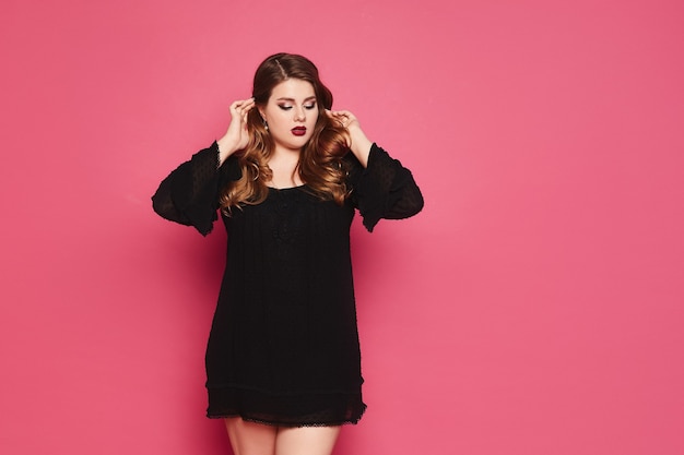 Menina modelo plus size com maquiagem brilhante e lábios carnudos e vermelhos em um vestido preto curto corrige o cabelo contra um fundo rosa.