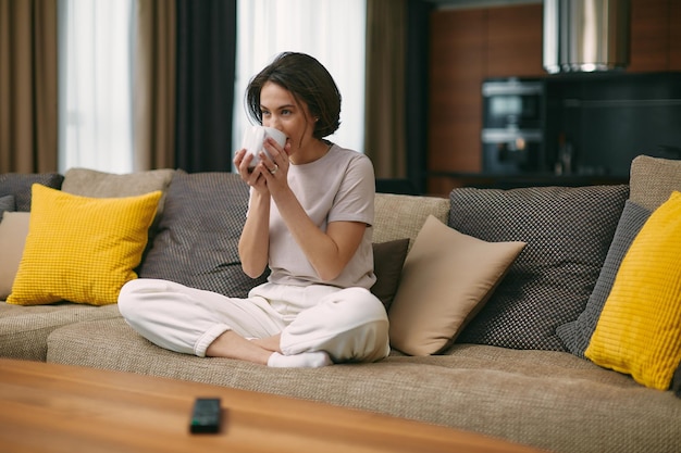 Menina moderna bebendo café ou chá segurando caneca sentada no confortável sofá assistindo filme da série de tv