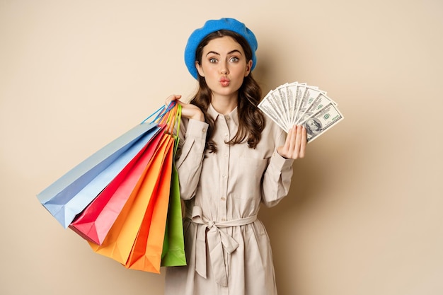 Menina moderna elegante sentindo satisfação ao fazer compras, posando com dólares de dinheiro e sacolas de compras, fundo de estúdio bege.