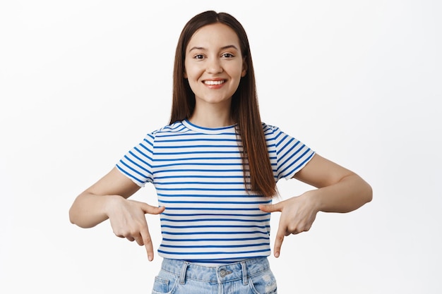 Menina morena alegre apontando os dedos para baixo, mostrando maquete, espaço vazio de anúncio abaixo, convidando para visitar o local, confira informações de venda, de pé contra um fundo branco e sorrindo satisfeito