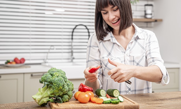 Menina morena alegre corta legumes na salada no fundo do interior da cozinha moderna.