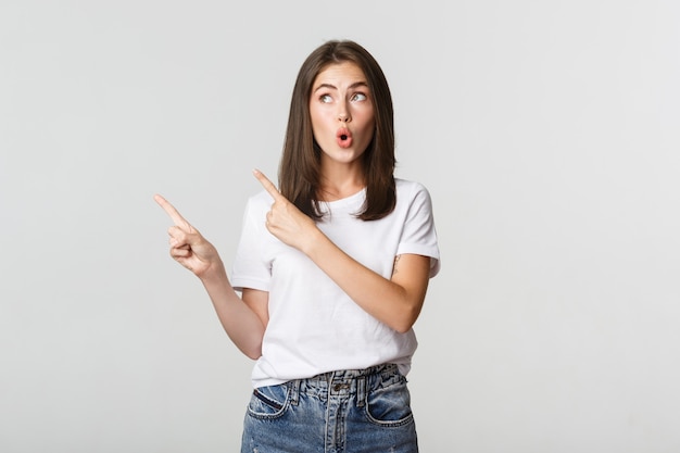 Menina morena atraente animada em t-shirt branca apontando os dedos no canto superior esquerdo, parecendo curiosa.