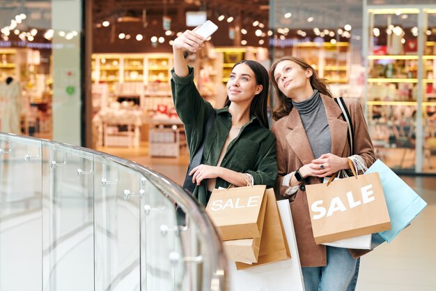 Menina morena atraente com camisa de veludo segurando sacolas de compras e fotografando com um amigo no shopping