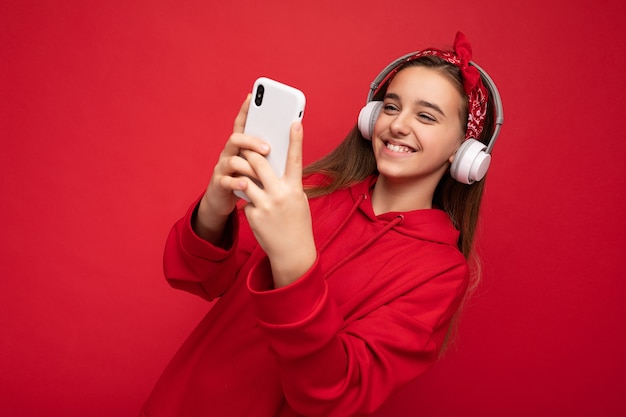 Menina morena atraente positiva vestindo um capuz vermelho isolado em um fundo vermelho segurando e usando