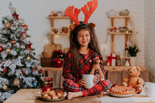 Menina morena bonitinha de pijama vermelho quadriculado com chifres de rena na cabeça está comendo um bolo de natal e bebendo chá de uma caneca branca em uma cozinha lindamente decorada. estilo de vida, hygge