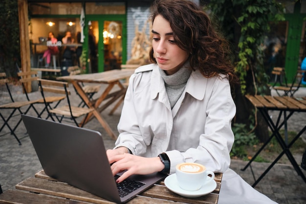 Menina morena casual atraente em trench coat estudando com confiança no laptop com café no café ao ar livre