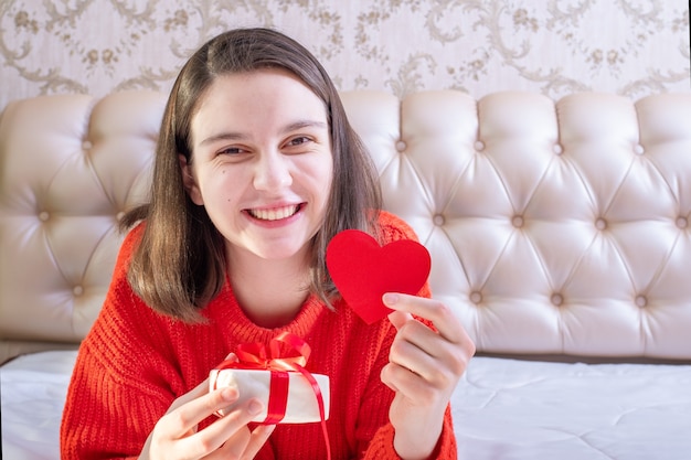 Menina morena caucasiana sorridente feliz mostrando uma caixa de presente de natal com fita e coração de formato vermelho de papel