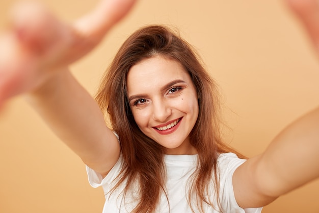 Menina morena com cabelos longos, vestida com uma camiseta branca faz uma selfie sobre o fundo bege no estúdio.