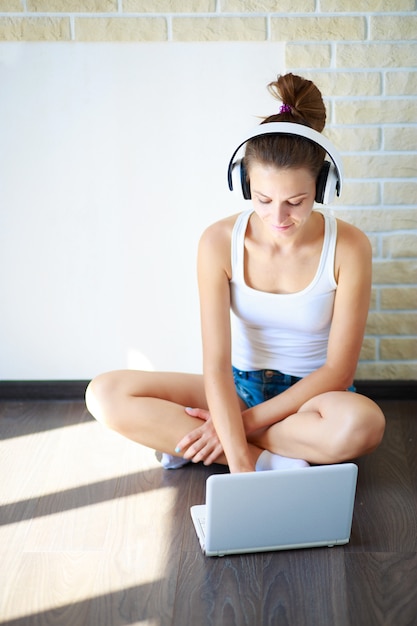 menina morena com fones de ouvido, ouvindo música em um laptop em uma sala vazia