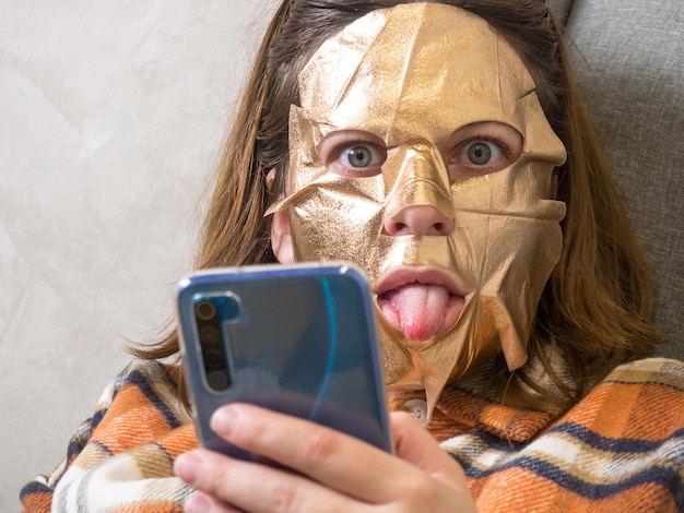 Menina morena com um telefone celular em uma máscara de folha de ouro no rosto. relaxe em uma poltrona durante tratamentos de beleza em casa em uma poltrona.
