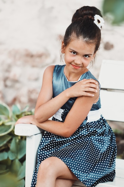 Menina morena com um vestido azul com bolinhas brancas se senta em um banco no verão. Foto de alta qualidade