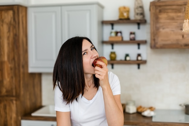 Menina morena com uma camiseta branca na cozinha come uma maçã.