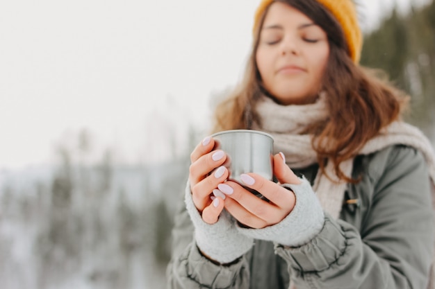 Menina morena de chapéu de malha amarelo com caneca de metal de chá quente na floresta ao ar livre no inverno, foco seletivo