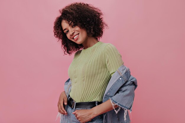 Menina morena de top verde de ótimo humor posando em fundo rosa Muito jovem na jaqueta jeans olha para a câmera no pano de fundo isolado