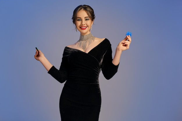 Menina morena de vestido preto e joias brilhantes. Sorrindo, mostrando duas fichas azuis e verdes, posando em fundo roxo. Pôquer, cassino. Fechar-se