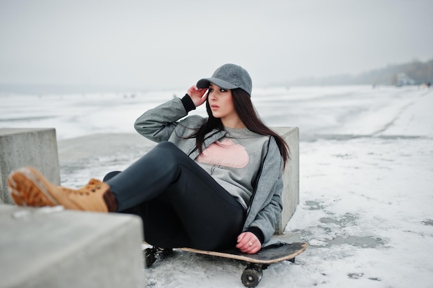 Menina morena elegante no estilo de rua casual de boné cinza com prancha de skate no dia de inverno