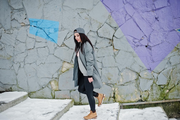 Menina morena elegante no estilo de rua casual de boné cinza no dia de inverno contra a parede colorida