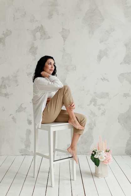 Menina morena elegante sentada em uma cadeira alta branca com um buquê de flores no chão