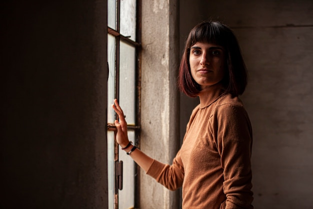 Menina morena em frente a uma janela, olhando para a câmera, imagem com pouca luz