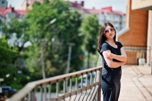 Menina morena em óculos de sol vestido preto posando na rua da cidade