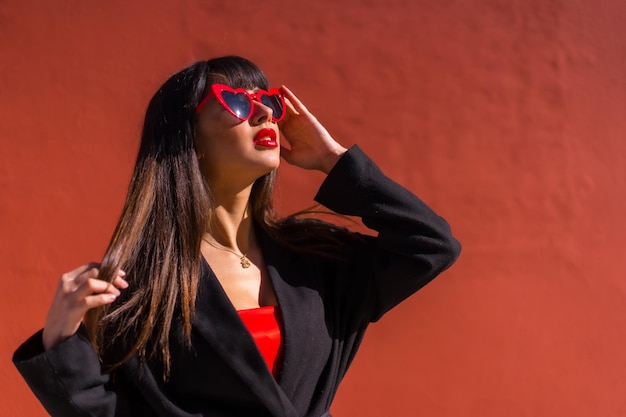 Menina morena em um retrato em um fundo vermelho com óculos de corações pose de moda do dia dos namorados