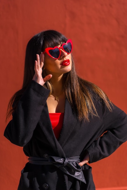 Menina morena em um retrato em um fundo vermelho com óculos de corações pose de moda do dia dos namorados