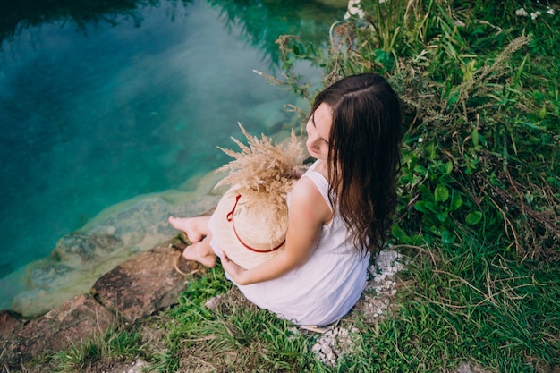 Menina morena em um vestido branco e segurando um chapéu de palha perto do lago com um buquê de flores.