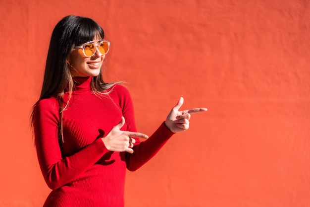 Menina morena em um vestido laranja primavera e óculos de sol, apontando para uma oferta à direita. Copie e cole o espaço. Caucasiano muito um fundo vermelho