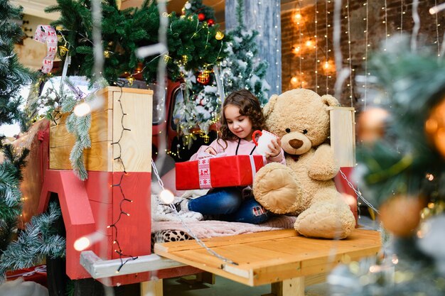 Menina morena encaracolada sentada perto de árvore decorada de natal e ano novo com um grande ursinho de pelúcia, xícara e abra a caixa vermelha de presente