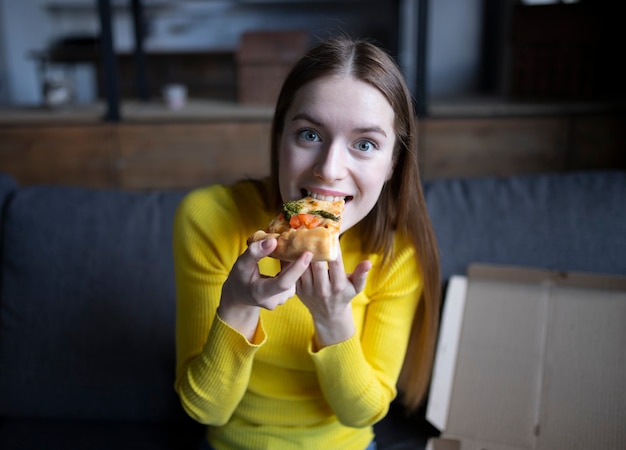 Menina morena engraçada com suéter amarelo comendo pizza em casa