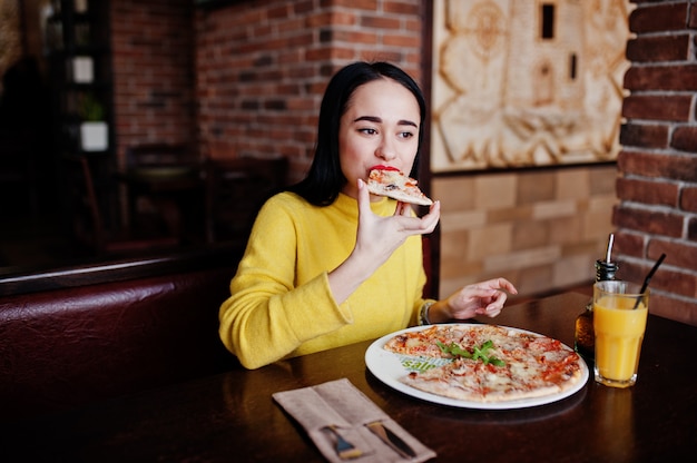 Foto menina morena engraçada na camisola amarela que come a pizza no restaurante.