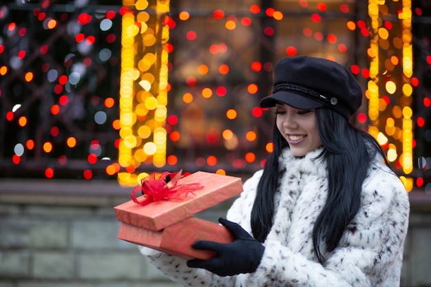 Menina morena feliz usando boné da moda, segurando a caixa de presente com fita e arco, posando para a rua. Espaço para texto