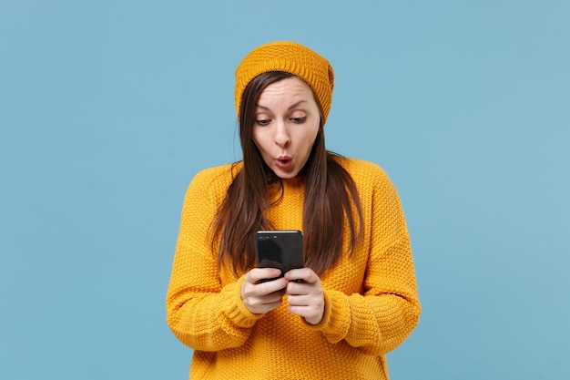 Menina morena jovem chocada na camisola amarela e chapéu posando isolado no retrato de estúdio de fundo azul. Conceito de estilo de vida de pessoas. Mock up espaço de cópia. Usando o telefone celular, digitando mensagem sms.