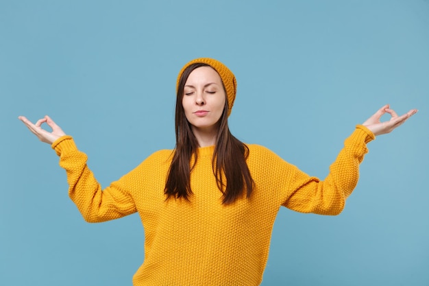 Menina morena jovem de suéter amarelo e chapéu posando isolado no retrato de estúdio de fundo azul. Conceito de estilo de vida de pessoas. Mock up espaço de cópia. Segure as mãos em gesto de ioga, relaxando meditando.