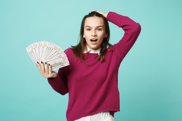 Menina morena jovem em roupas casuais posando isolado no retrato de estúdio de fundo azul. Conceito de estilo de vida de emoções de pessoas. Espaço de cópia simulado Segure na mão o ventilador de dinheiro em notas de dólar