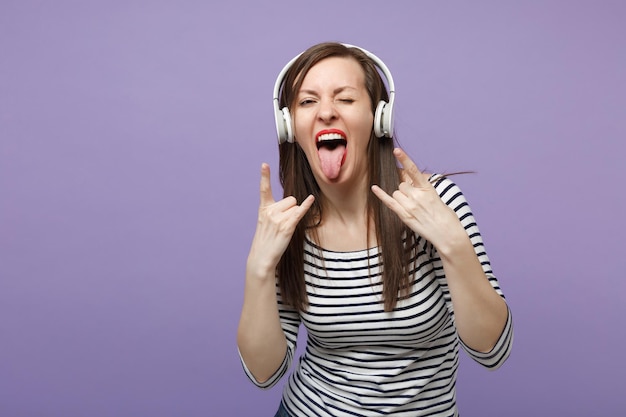 Menina morena jovem em roupas listradas casuais posando isolado no retrato de estúdio de fundo roxo violeta. conceito de estilo de vida de emoções sinceras de pessoas mock up copy space ouça música em fones de ouvido