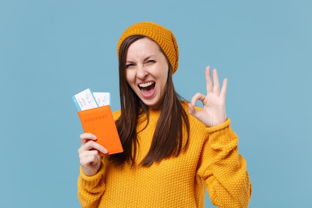 Menina morena jovem engraçada no suéter chapéu posando isolado no retrato de estúdio de fundo azul. Conceito de estilo de vida de pessoas. Mock up espaço de cópia. Segure os bilhetes de embarque do passaporte mostrando o gesto OK.