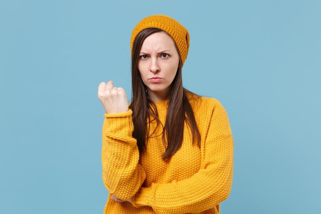 Menina morena jovem estrita com raiva na camisola amarela e chapéu posando isolado no retrato de estúdio de fundo de parede azul. Conceito de estilo de vida de emoções sinceras de pessoas. Mock up espaço de cópia. Apertando o punho.